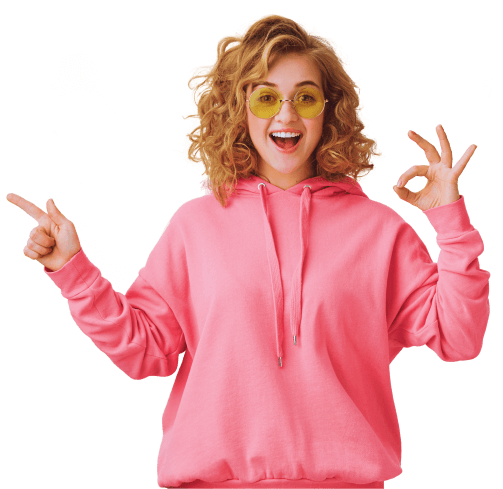 Jeune femme enthousiaste aux cheveux bouclés portant des lunettes de soleil jaunes et un sweat à capuche rose, faisant le signe 'OK' avec une main et pointant de l'autre, exprimant la positivité et l'amusement